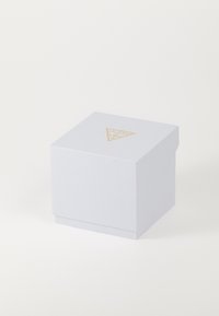 Boîte en carton blanc texturé, de forme carrée, comportant un logo en triangle doré avec le texte "GUESS?" en dessus. Design minimaliste, aucun matériel visible.