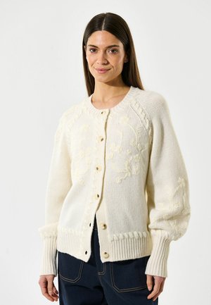 Krämfärgad stickad kofta med broderade blommönster, ribbade ärmslut och knappar nedtill framtill; avslappnad passform med puffärmar.