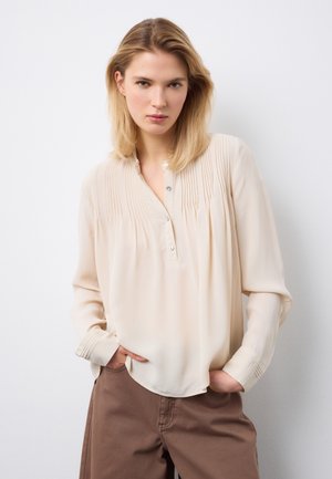 Blonde Frau, die eine cremefarbene, plissierte Bluse mit langen Ärmeln und braune Hosen trägt, mit den Händen in den Taschen, vor einem weißen Hintergrund stehend.