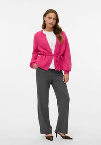 Cardigan tricoté rose vif avec une ceinture à nouer, un col rond et des manches amples, associé à un pantalon gris et des escarpins à bout pointu.