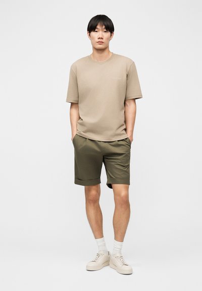 Νεαρός άνδρας όρθιος με τα χέρια στις τσέπες, φοράει μπεζ μπλουζάκι, σορτς λαδί, λευκές κάλτσες και μπεζ αθλητικά παπούτσια σε απλό φόντο.