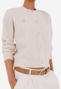 Cardigan lavorato a maglia beige chiaro con maniche lunghe, orlo a coste e bottoni decorativi. Presenta due tasche frontali con dettagli testurizzati.