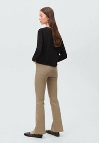 Črna majica z dolgimi rokavi, svetlo rjave široke kavbojke in črne balerinke s bodicami. Lase spuščene, obrnjen rahlo levo, minimalistična ozadje.