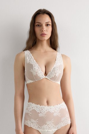 Jeune femme debout contre un mur blanc, portant un soutien-gorge en dentelle beige et une culotte taille haute assortie en dentelle.