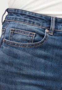 Gros plan d'une poche de jean en denim bleu avec couture visible, rivets métalliques et passant de ceinture sur un fond en tissu blanc.
