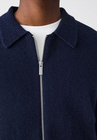 Granatowa kurtka na zamek z teksturowanego materiału, z ostrym kołnierzem i srebrnym metalowym zamkiem, noszona na białej koszulce z okrągłym dekoltem.