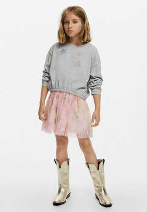 Grauer Sweatshirt mit Sternenverzierungen, pinker Tüllrock mit floralem Muster und glänzenden goldenen Cowboystiefeln. Einfaches, lässiges Outfit.
