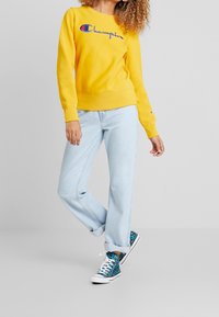 Felpa gialla in cotone con logo "Champion" ricamato, abbinata a jeans a gamba larga azzurri e sneakers con una vivace stampa leopardata.