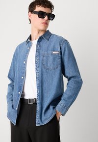 Denim overhemd in lichtblauw, met knoopsluiting en een borstzak. Draagt over een witte t-shirt, gecombineerd met zwarte broek en zonnebril.