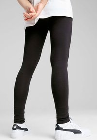 Persona vestida con leggings negros, calcetines blancos y zapatillas Puma blancas con detalles en negro, de pie con las manos entrelazadas detrás de la espalda.