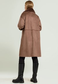 Brown faux fur coat with a wide collar and dropped shoulders, featuring a textured finish and a straight silhouette. Black knee-high boots visible.