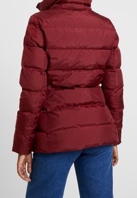 Vinröd vadderad jacka med hög krage, sydda paneler och en figursydd siluett, med en slät, glänsande yta.