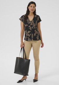 Blusa nera a fiori con scollo a V e maniche corte, abbinata a pantaloni chiari attillati. In una mano è tenuta una grande borsa a tracolla nera con manici lunghi.