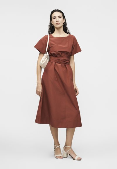 Γυναίκα με μεσαίου μήκους φόρεμα σε σκουριά απόχρωση και κοντά μανίκια, που κρατά μια μικρή μπεζ τσάντα ώμου και φοράει μπεζ πέδιλα με τακούνι.