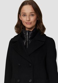 Schwarzer Wollmischung Doppelreiher-Mantel mit großen Revers und Fronttaschen, über einer schwarzen Daunenjacke mit hohem Kragen und Reißverschluss getragen.