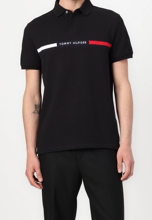 Mężczyzna ubrany w czarną koszulkę polo z krótkim rękawem z logo Tommy Hilfiger oraz czerwonymi i białymi poziomymi paskami na piersi.