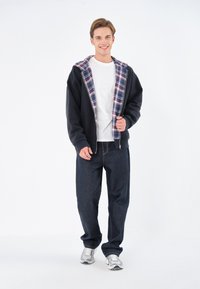 Sudadera negra con cremallera y forro a cuadros, combinada con una camiseta blanca y jeans de mezclilla oscura. Modelo llevando zapatillas plateadas.