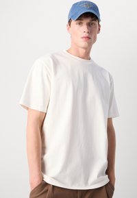 Balta trumpa rankovė t-shirt, pagaminta iš medvilnės, su apvaliu kaklu. Dėvima su šviesiai mėlyna kepuraite, su geltonai išsiuvinėtu tekstu.