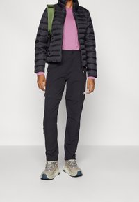 Sort oppustelig jakke med høj krave, lyserødt langærmet skjorte og sorte bukser med lynlås. Beige vandrestøvler med lyserøde snørebånd. Grøn rygsækrem.