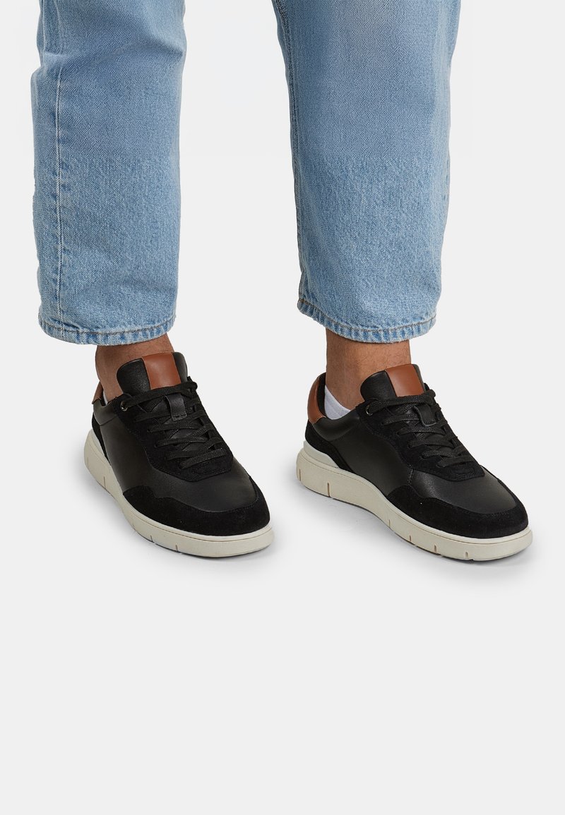 Sneakers neri con tomaia in suede e pelle. Presentano un accento in pelle marrone sul tallone e una suola in gomma beige. Indossati con jeans azzurri chiari.