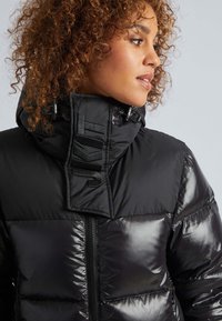 Chaqueta acolchada negra con cuello alto, textura brillante, prominente marca "JKIT" en el cuello y cierre de cremallera.