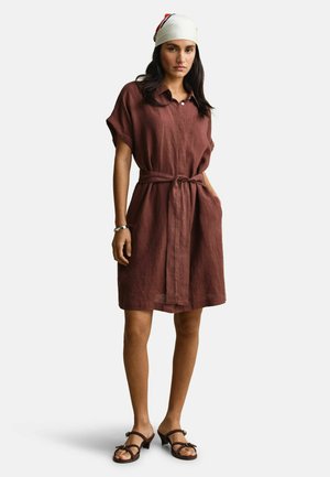 Frau trägt ein braunes, kurzärmeliges Kleid mit Gürtel, ein weißes Kopftuch mit roten und blauen Streifen, schwarze Sandalen mit Absatz und ein silbernes Armband.