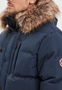Marineblaue Steppjacke mit Kunstfellkapuze, gestepptem Design, verstellbaren Kordeln und Logo-Patch am Ärmel.