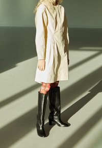 Vestido de color crema con textura y mangas largas, combinado con leggings rojos estampados y botas de cuero negro hasta la rodilla.