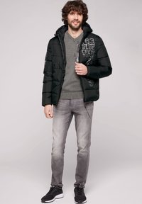 Chaleco acolchado negro con capucha y cremallera, que presenta bolsillos laterales y un detalle de logo en el pecho. Combinado con un suéter gris y jeans gris claro.