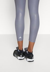 Nohy v lesklých šedých zkrácených legínách, černých ponožkách Adidas a černobílých teniskách Adidas, stojící na neutrálním pozadí.