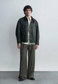 Bărbat tânăr purtând ochelari, jachetă închisă la culoare verde, cămașă albă, pantaloni gri largi și pantofi negri, stând în fața unui fundal simplu.