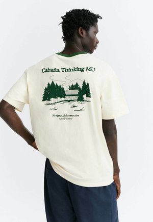 Homme portant un t-shirt crème avec un graphisme d'une cabane verte et des arbres et le texte "Cabaña Thinking MU" dans le dos, debout sur fond blanc.