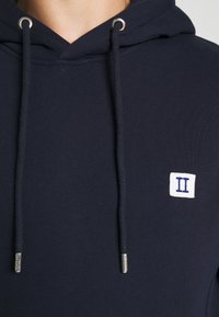 Sweat à capuche bleu marine avec cordons et une petite pièce blanche affichant un chiffre romain II bleu sur la poitrine.