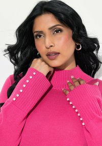 Pull en tricot fuchsia avec un col montant, des poignets côtelés embellis de perles blanches, et une texture lisse. Gros plan sur des mains sur le tissu.