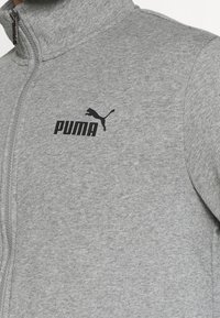 Šedá zipová bunda vyrobená z měkkého materiálu. Má vysoký límec a černé logo Puma na levé části hrudi.