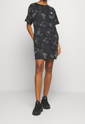 Person trägt ein locker sitzendes schwarz-grau camouflagegemustertes Kleid mit kurzen Ärmeln und schwarze CAT-Sneaker vor einem einfarbigen weißen Hintergrund.