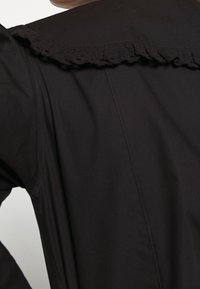 Manteau noir avec un col en dentelle froncée, texture lisse et design structuré. Vue arrière montrant des détails de couture visibles.
