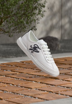 Sneaker bianca con logo decorativo nero sul lato, lacci bianchi, che fluttano sopra una superficie esterna piastrellata in legno e pietra.