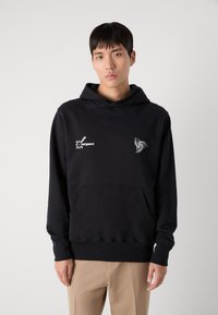 Hoodie noir avec une poche kangourou, arborant des impressions graphiques blanches sur la poitrine gauche et la manche. Fabriqué en tissu doux et texturé.