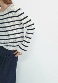 Persoon draagt een wit en marineblauw gestreepte gebreide trui met lange mouwen en marineblauwe broek, staand met één hand in de zak tegen een effen muur.