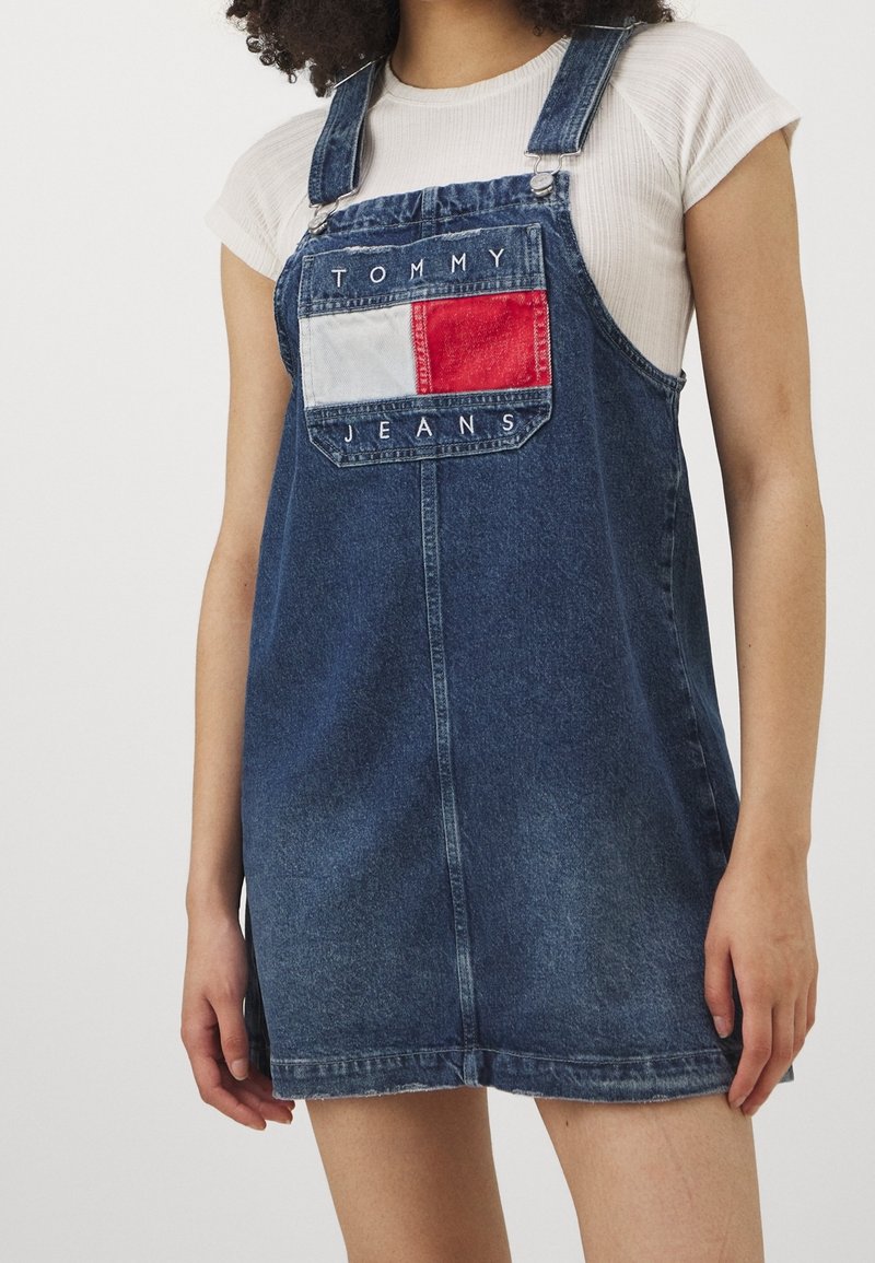 Denimová šaty s kšandami, přední kapsou s červenou a bílou obdélníkovou nášivkou a vyšitým nápisem "TOMMY JEANS" v bílé barvě.