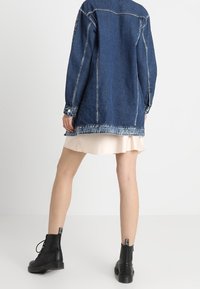 Džínová bunda s volným střihem, tmavě modré barvy a bílým šitím. Kombinována se světle béžovou sukní a černými kotníkovými botami.