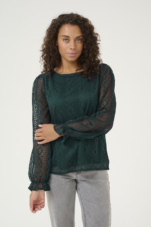 Dunkelgrüne Spitzenbluse mit geometrischem Muster, langen durchsichtigen Puffärmeln mit Rüschenmanschetten und rundem Ausschnitt. Getragen mit hellgrauen Jeans.
