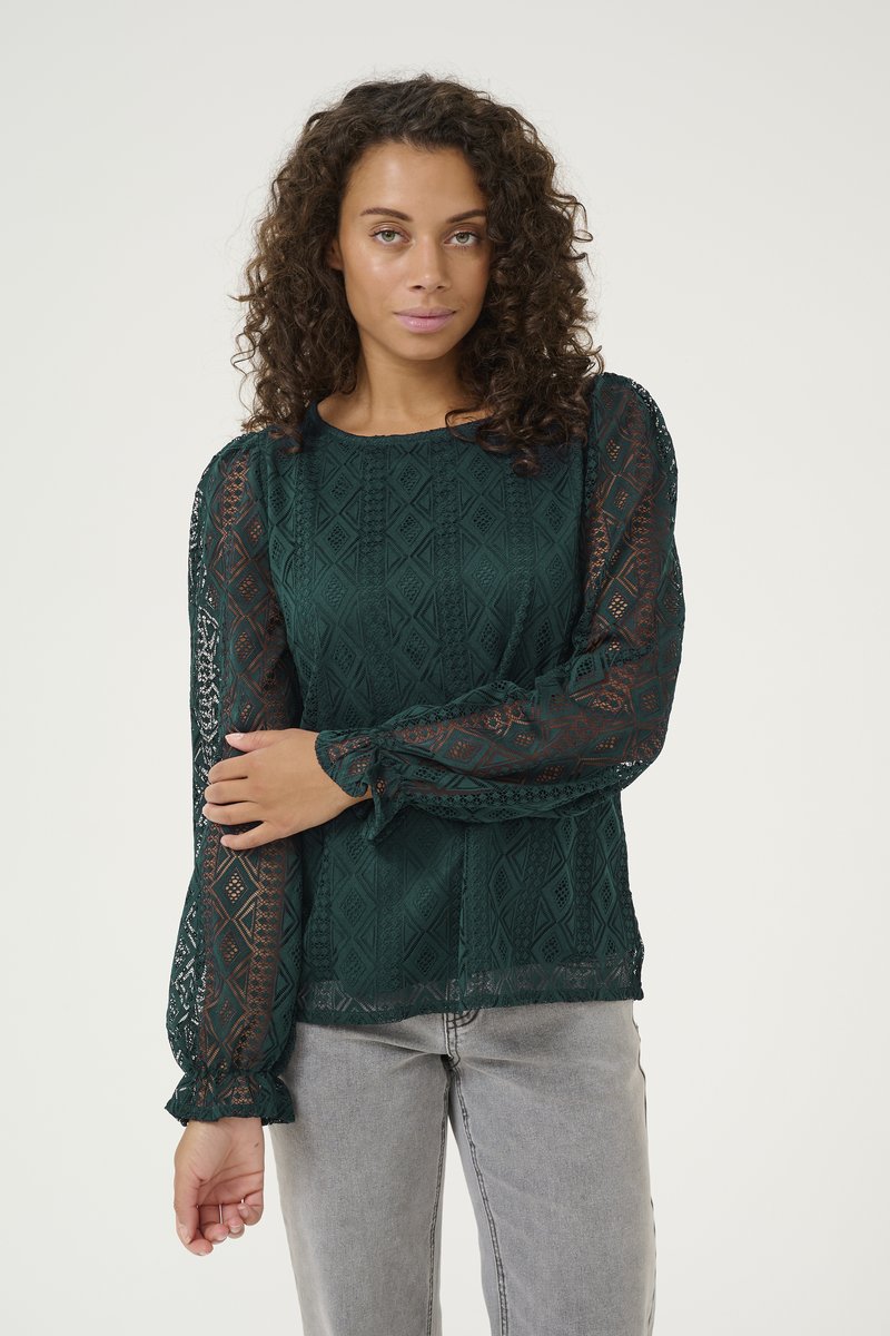 Blusa in pizzo verde scuro con motivo geometrico, maniche lunghe e trasparenti a sbuffo con polsini arricciati e scollatura rotonda. Indossata con jeans grigi chiari.
