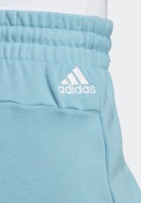 Světle modré tepláky Adidas mají elastický pas a bílé vyšívané logo. Tkanina vypadá měkce a hladce.