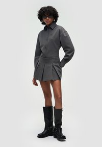 Camicia grigia con colletto abbinata a una mini gonna grigia plissettata. La modella indossa stivali neri fino al ginocchio e occhiali da sole.