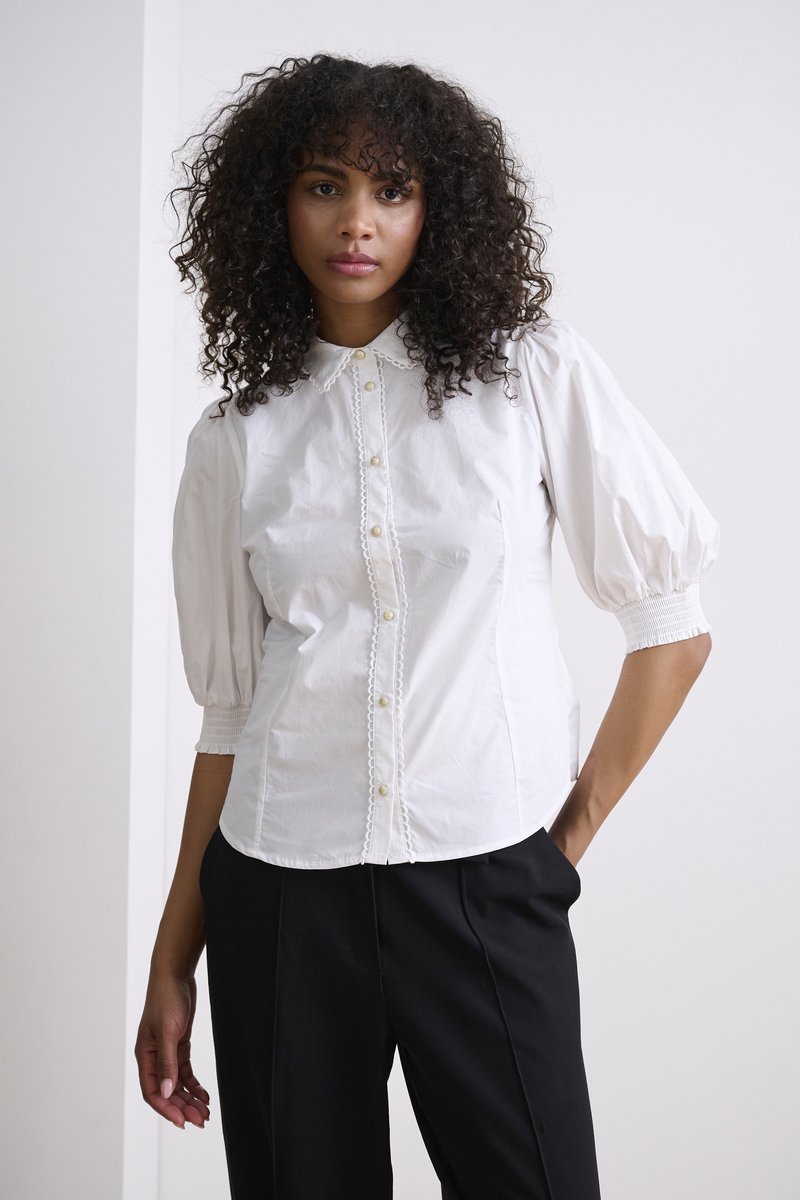 Blusa bianca con busto aderente, maniche a sbuffo e chiusura con bottoni. Presenta dettagli decorativi lungo la pattina e un tessuto morbido.