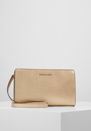 Gouden Michael Kors polsclutch met textuur oppervlak en afneembare riem op een wit tentoonstellingspodium.