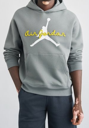 Personne portant un sweat à capuche gris Air Jordan avec un lettrage jaune et un logo Jumpman blanc, associé à un pantalon foncé, debout, les bras détendus.