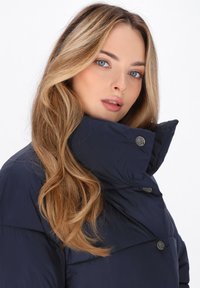 Cappotto puffer blu navy con colletto alto, bottoni a pressione, texture liscia e design fitted che si adatta al corpo.